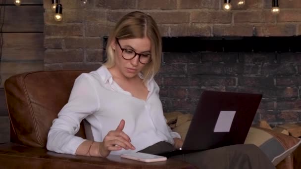 一位年轻的女商人 戴着眼镜 用笔记本电脑和智能手机往下看 在一个明亮的大办公室里坐在椅子上做生意 — 图库视频影像