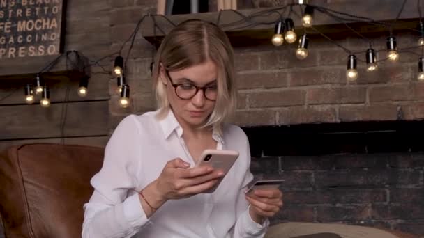 若いビジネス女性がオンラインで購入する大きな明るい部屋で椅子に座ってスマートフォンやクレジットカードを使用します — ストック動画