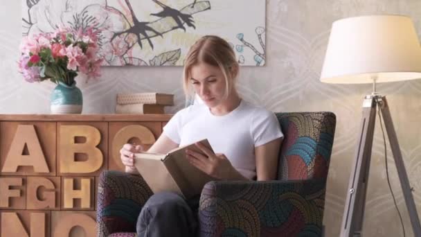 美しい若い女性は 自宅のリビングルームで椅子に横たわってリラックスした読書フィクションの本をお楽しみください 可愛いですティーン女の子休みますオン — ストック動画