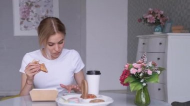 Genç bir sarışın kadın haberleri Smartphone 'da okuyor. Hafif bir kafede bir sandalyede oturuyor, bir kadın kahve içiyor ve öğle yemeği molasında sandviç yiyor.