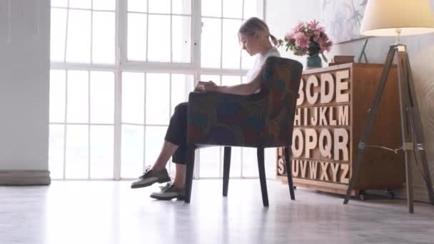 明るい部屋の家で椅子に座って文学を勉強している本を読んでいる若いブロンドの女性の側面図 — ストック動画
