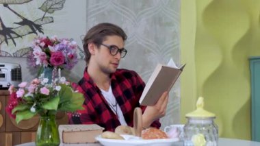 Modern ferah mutfakta masada oturan, kitap okuyan, kahve içen, kitabı kapayan ve pencereden dikkatlice bakan beyaz bir adamın portresi.