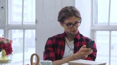 Parlak bir odada sandalyede oturan genç bir adam akıllı bir telefon kullanarak haberleri seyrediyor.