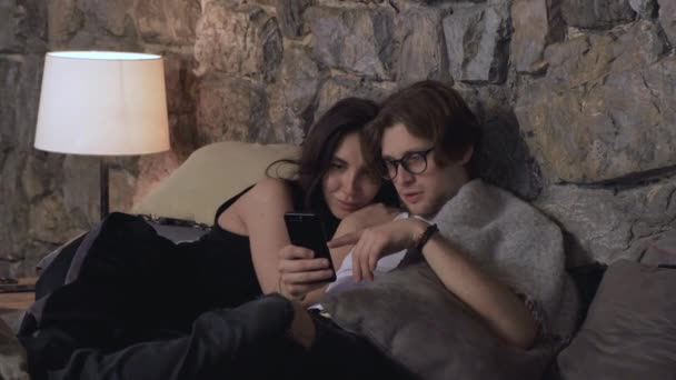 若いカップルがベッドで寝そべっている暗い部屋の家にいるスマートフォンの写真を見てチャット — ストック動画