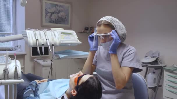 一个年轻的牙科医生在眼镜上放了一个镜子和计算机 准备检查一个病人的白衣和面罩 — 图库视频影像