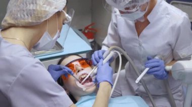 Hasta ile çalışan, dişlerini fırçalayan ve hasta, beyaz önlük ve maskeyi inceleyen gözlüklü iki genç dişçinin yakın çekimi.