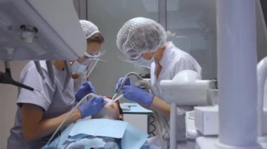 Gözlüklü iki genç dişçi hastanın diş temizliği ve muayenesi için çalışıyor. Hasta, beyaz önlük ve maske.