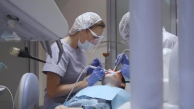 Bir dişçi ofisinde hareketli bir çekim, genç bir kadın iki dişçi hastaların dişlerini muayene ediyor ve üzerinde çalışıyor..