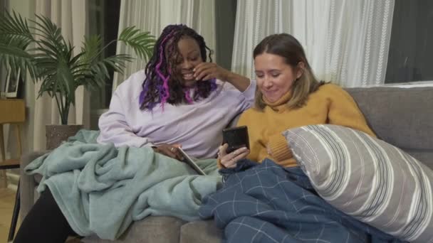 来自不同民族的非洲裔美国人和欧洲裔年轻女性的朋友们看着智能手机里的照片 呆在家里 舒适的公寓里 — 图库视频影像