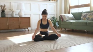 Evdeki oturma odasında gerinen genç bir kadın. Genç Hintli kadın meditasyon yapıyor. Halıya oturuyor ve yoga yapıyor.