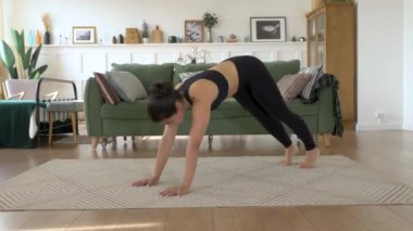 Hintli kadın, evde yoga yaparken köpek pozuna doğru eğiliyor.