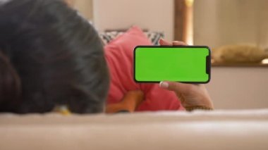 Oturma odasında yeşil ekranlı akıllı telefon kullanan Hintli kız filmi izliyor, video içeriği. Evde Kanepeye Uzanan Genç Kadın. İzleme cihazları. Yatay pozisyonda akıllı telefon var. İçeride. Yakın çekim.