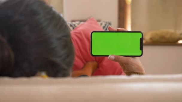 リビングルームで緑色の画面を持つスマートフォンを使用してインドの女の子映画 ビデオコンテンツを見て 自宅でソファに寝そべっている若い女性 マーカーを追跡中 水平位置にスマートフォン 室内だ — ストック動画