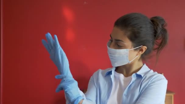 マスクを身に着けている若いインドのシリアル女性は居心地の良い明るい部屋で医療用シリコーンゴム手袋を置きます — ストック動画