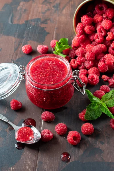 fresh raspberry jam in a glass jar on a wooden table, next to fresh raspberries and mint leaves. concept of homemade jam, preparation for winter,  selective focus and copy space