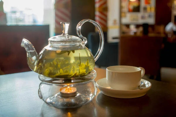 transparent teapot with green hot tea and a cup on a wooden table