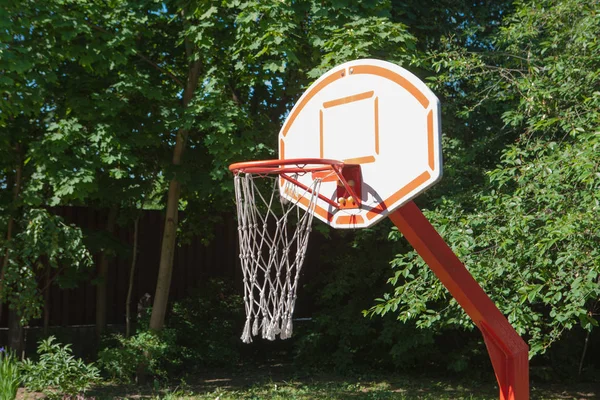 Red white basketball hoop on a sunny day. with rope mesh on a background of green trees — Stock Photo, Image