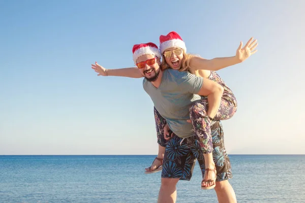 Homme piggyback à sa petite amie dans chapeaux de Noël et lunettes de soleil à la plage de la mer — Photo