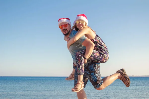 Noel şapka ve güneş gözlüğü plajda denizin arkadaşına piggybacking adam