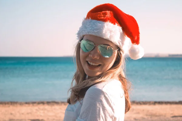 Porträt einer hübschen jungen Frau mit Weihnachtsmann-Hut und Sonnenbrille an einem sonnigen Strand. gemildert. — Stockfoto