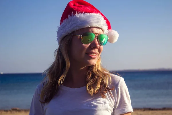 Santa Claus şapka ve güneş gözlüğü güneşli bir plaj oldukça genç bir kadın portresi. Tonda.