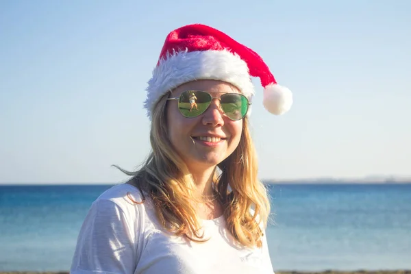 Portrait d'une jolie jeune femme en chapeau de Père Noël et lunettes de soleil sur une plage ensoleillée. tonique . — Photo