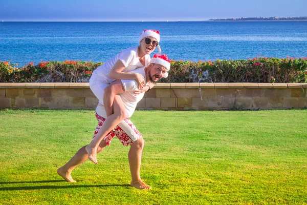 Homme piggyback à sa petite amie dans chapeaux de Noël et lunettes de soleil à la plage de la mer — Photo