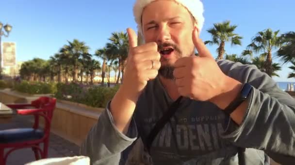 Porträtt av positiva man i jul hatt på beach café med palmer och havet — Stockvideo