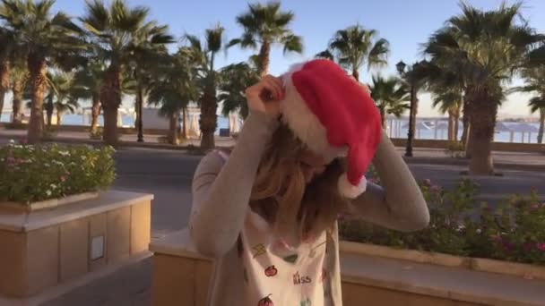 Портрет молодой и красивой девушки в рождественской шляпе в кафе с пальмами и морем — стоковое видео