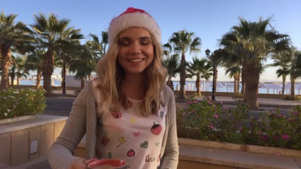Portré, fiatal és gyönyörű nő kávéfogyasztás karácsonyi kalapban. pálmák — Stock videók