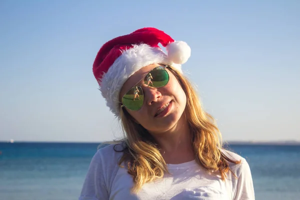 Portrait of a pretty young woman in Santa Claus hat and sunglasses on a sunny beach. Toned. — Stock Photo, Image