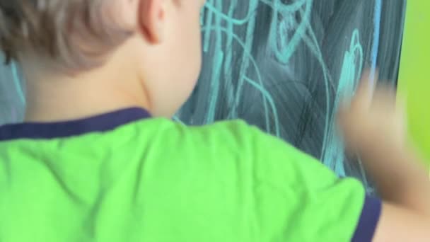 3 yaşındaki yeşil tişörtlü çocuk kara tahtaya tebeşir çiziyor. — Stok video