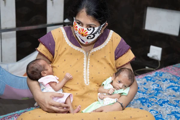 Young Mother Wearing Mask sitting on bed with Her Two Newborn Twin Babies, Stay Home Taking Extra Care Due To Covid-19 Pandemic. Indian Mother With Her Babies.