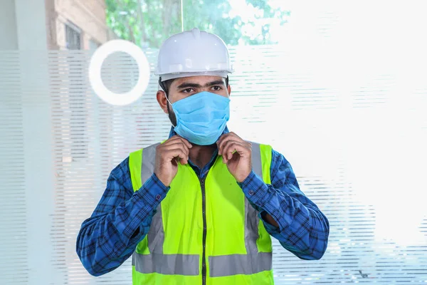 Maske takan genç mühendis gülümsüyor, sakallı adama yakın çekim sarı yelek ve beyaz kasklı mavi gömlek giyiyor, tecrit bittikten sonra işe dönüyor.