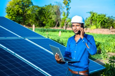 Genç erkek mühendis elinde dizüstü bilgisayarla telefonda konuşuyor. Güneş panellerinin yanında duruyor. Temiz mavi gökyüzü arka planına sahip tarım arazisi, yenilenebilir enerji, temiz enerji..
