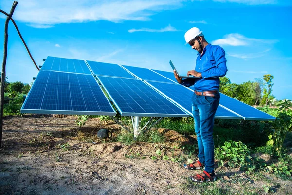 Dizüstü bilgisayarı olan genç erkek mühendis güneş panellerinin yanında duruyor. Tarım arazisi açık mavi gökyüzü arka planı, yenilenebilir enerji, temiz enerji..