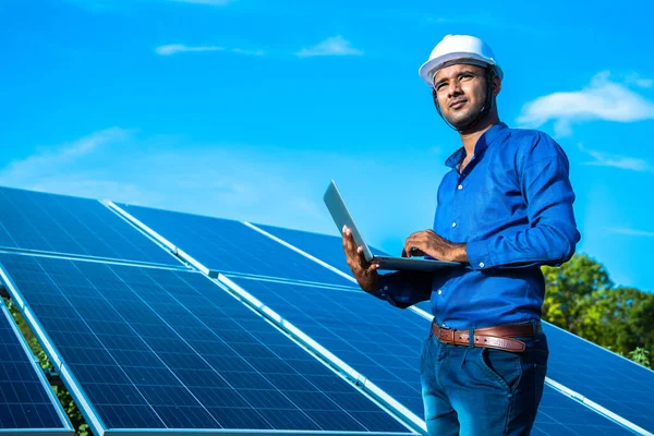 Dizüstü bilgisayarı olan genç erkek mühendis güneş panellerinin yanında duruyor. Tarım arazisi açık mavi gökyüzü arka planı, yenilenebilir enerji, temiz enerji..
