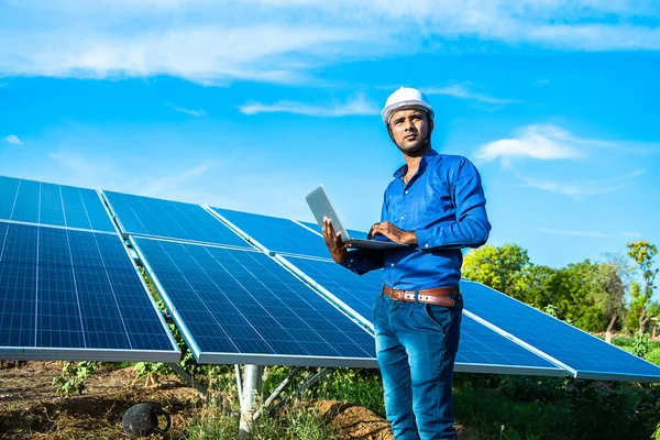 Dizüstü bilgisayarı olan genç erkek mühendis güneş panellerinin yanında duruyor. Tarım arazisi açık mavi gökyüzü arka planı, yenilenebilir enerji, temiz enerji..