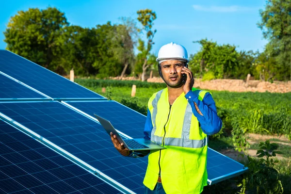 young male technician worker wearing green vests and helmet with laptop in hand talking on phone about the maintenance and installation of the solar panels standing in field, technology in agriculture