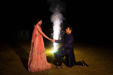 Hintli bir adam dizlerinin üzerine çöküp kız arkadaşının el teklifini geleneksel kıyafetle tutarak arka planda havai fişeklerle bayram kutluyor. Romantik teklif.