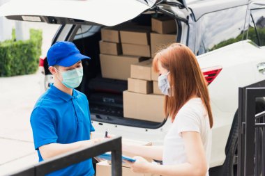 Asyalı genç kurye posta kutusuyla çevrimiçi alışveriş yapıyor üniformalı koruyucu yüz maskeli Coronavirus. Evdeki kağıt defter üzerinde kalem imzası kullanan servis elemanı müşteri yapıyor, COVID-19.