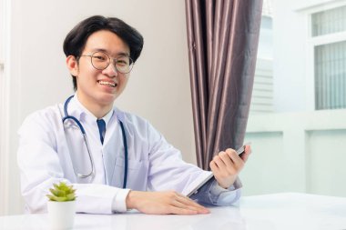 Mutlu Asyalı genç doktor yakışıklı adam gülümsüyor ev ofisinden çalışma gözlükleri takıyor, hastane ofisindeki siyah modern dijital tablet bilgisayar kullanıyor, Teknoloji sağlık ve tıp konseptini kullanıyor.