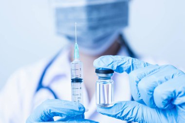 Asyalı kadın doktor ya da hemşire üniformalı ve steteskop takan eldiven takmış. Laboratuvarda elinde şırınga ve ilaç şişesi aşı şişesi, sağlık konsepti olan yüz maskesi takıyor.