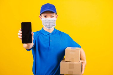 Asya 'lı genç teslimatçı erkek tişörtü ve şapka üniforması giyiyor. Yüz maskesi takıyor. Coronavirus ya da COVID-19 altında siyah akıllı cep telefonu ekranını gösteriyor.