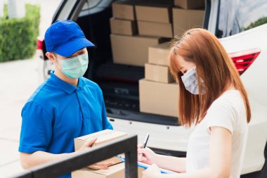 Asyalı genç kurye posta kutusuyla çevrimiçi alışveriş yapıyor üniformalı koruyucu yüz maskeli Coronavirus. Evdeki kağıt defter üzerinde kalem imzası kullanan servis elemanı müşteri yapıyor, COVID-19.