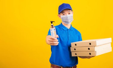 Asyalı yakışıklı teslimatçı yüz maskeli mikrop virüsü taşıyor. Elinde alkol dezenfekte edici jel ve kutular var. Stüdyo çekimi sarı arka planda izole edilmiş.