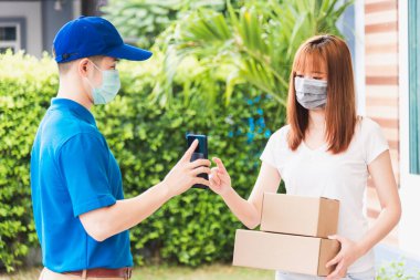 Asya teslimat ekspres kurye genç adam cep telefonundan kadın müşteri imzası için paket kutuları veriyor hem koruyucu yüz maskesi, sokağa çıkma yasağı altındaki kordemik koronavirüs COVID-19