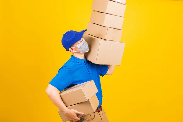 Asian young delivery worker man in blue t-shirt and cap uniform wearing face mask protective lifting stack a lot of boxes, under coronavirus or COVID-19, studio shot isolated yellow background