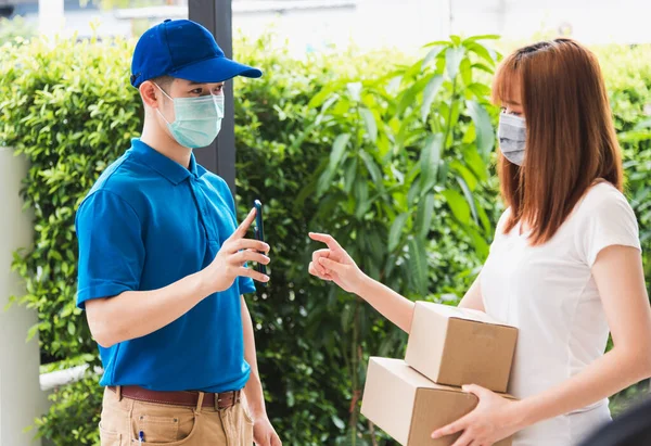 Asya teslimat ekspres kurye genç adam cep telefonundan kadın müşteri imzası için paket kutuları veriyor hem koruyucu yüz maskesi, sokağa çıkma yasağı altındaki kordemik koronavirüs COVID-19