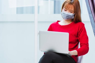 Asyalı genç iş kadını kırmızı tişört giyer yüz maskesi takar ev bilgisayarından çalışır Coronavirus hastalığını karantinaya alır veya COVID-19